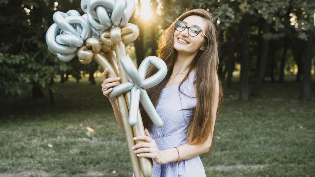    Девушка в очках с длинными волосами стоит и держит в руках букет из длинных шариков золотистого и серебристого цвета:Freepik