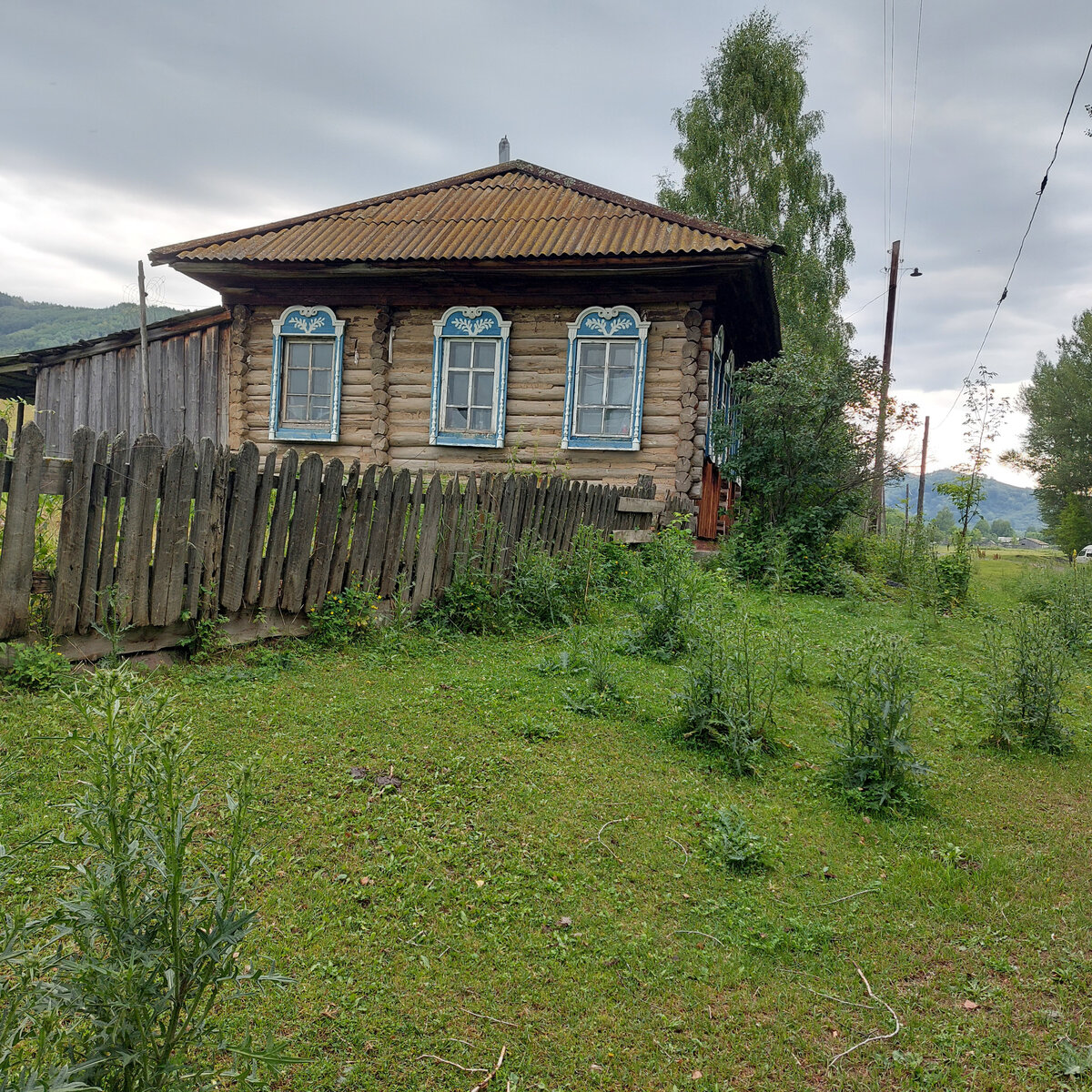 Красивейший дом в Усть-Тулатинке. Одно время здесь жил мой одноклассник ЛОБАНОВ НИКОЛАЙ. Чей дом сегодня-не знаю. Но слава Богу дом стоит и он в очень неплохом состоянии. Когда-то прямо под окнами шла дорожка в НИЗ. Сейчас все заросло травой и сегодня дорога идет там, где когда-то протекала протока от Тулаты, т.е. прямо по ее руслу. А НИЗ - так называется часть Усть-Тулатинки к Чарышу, т.е. вниз по течению ТУЛАТЫ. Я ходила тут часто в бабушке и дяде Саше в самый низ деревни к Камешку. Там и стоял почти точно такой же большой дом Иунихиных. С такими же красивыми резными наличниками. Только тогда их почему-то не красили так ярко... Одну половину у родителей бабушки отобрали при раскулачивании, отдали Лобанову Егору МАрковеевичу и от их фляги с керосином в сенях дом и сгорел... Это еще одна из трагедий нашей семьи... Но я помню запах перги в том доме, запах ульев (стояли в подвале), широченные гладкие доски пола в доме, в сенях, и на крыльце... Дом был уже тогда темного цвета. Возможно это была лиственница... Сени были сделаны их толстенных и широченных довольно гладких досок без щелей... Пазов даже почти не было... Окошко в сенях  было маленькое и высокое...Само место было у реки, с садом и мягчайшим огородом...В этом доме, кстати, тоже жило две семьи ( в моем детстве). Похоже, КРАСНАЯ ВЛАСТЬ уплотнила тогда зажиточных селян, разделив их дома на две семьи, а часть подобных  прекрасных домов ведь тогда была просто разобрана и вывезена НА НУЖДЫ СОВЕТСКОЙ ВЛАСТИ. Парадокс: дед мой боролся за Советскую власть, погиб, а его жену и пятерых детей не пожалели и выгнали из их же половины дома!!! Так что у бабушки с обеих сторон были у родителей подобные, а может и чуть больше, прекрасные дома и воооот.ни одного нет...А тут на фото, если посмотреть справа, то там уже виден косогор ВЕРХА деревни.