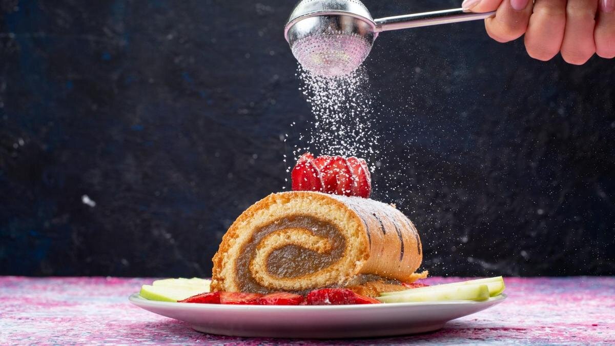    Рулет лежит на тарелке и его посыпают сахарной пудрой из ситечка:Freepik