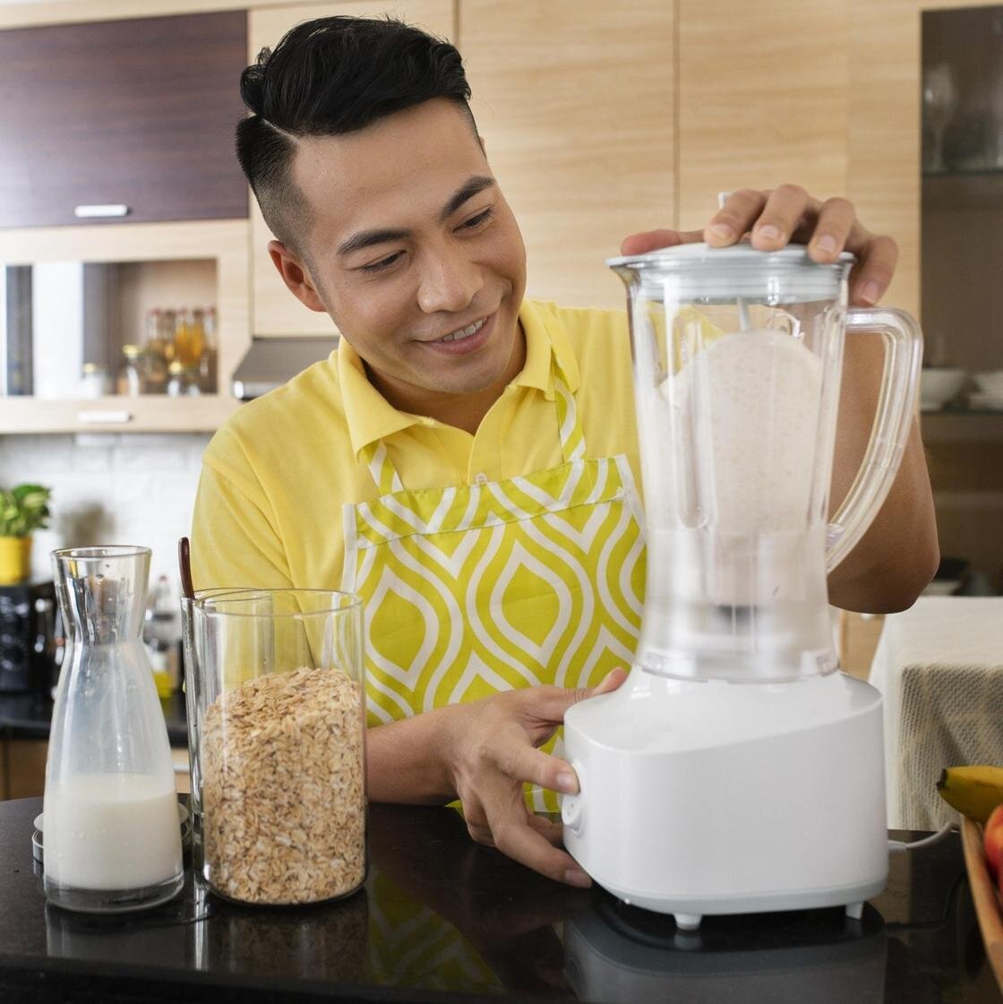    Молодой парень взбивает в блендере сахарную пудру из сахара. Рядом на столе стоят стеклянные емкости с молоком и овсяными хлопьями:Freepik