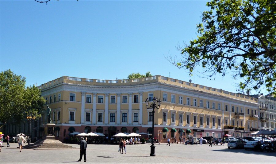  1. На полукруглой площади в центре Приморского бульвара, где находится памятник Дюку де Ришелье, по проекту известного петербургского архитектора Авраама Мельникова в 1827 – 30-х годах были сооружены два полуциркульных здания. Одно из этих зданий (по Приморскому бульвару, 7) предназначалось сначала для «присутственных мест», где располагались в свое время Одесская городская дума, приказ общественного призрения, строительный комитет, коммерческий суд и прочие административные учреждения. Именно в этом здании 4 октября 1916 года царский военный трибунал приговорил будущего одиозного комбрига Рабоче-крестьянской Красной армии Григория Котовского за ре­волюционную деятельность к смертной казни через повешение. Второе здание  (по Примор­скому бульвару, 8) первоначально при­надлежало частному лицу, а в 1840-х го­дах было превращено в фешенебельную «Санкт-Петербургскую гостиницу». Здесь часто останавливались известные люди того времени для отдыха, а кто-то и для поправки здоровья в Южной Пальмире. Расположенные на Приморском буль­варе против памятника Ришелье, эти здания в совокупности с Потемкинской лестницей образуют стройный архитектурный ансамбль, достойно представляя с морской стороны внешний фасад парадно-административного центра города.
Снимок сделан от одного из этих полуциркулярных зданий.