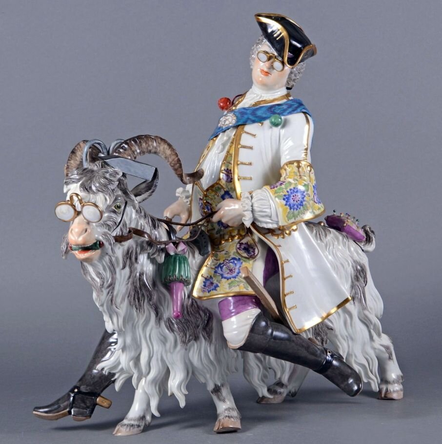 Здесь, скорее всего, вариант более поздний. И наряд сидящего на козле более богатый. Шляпа заменена треуголкой, расшитый золотом камзол, расстёгнутый на все пуговицы, рукава с широким обшлагом; ботфорты, как у высокого военного чина, со шпорами. И видимо, очень модное в те годы пенсне (как у всадника, так и у козла). Атрибуты портного - ножницы и утюг - повешены козлу на рога. Через плечо седока перекинута голубая орденская лента с условным овальным "орденом", где изображены ножницы, утюг и портновский метр. Такой же метр болтается сбоку вместо шпаги. У хвоста козла закреплена подушечка с воткнутыми в неё иголками.
"Портной на козле". Мейсен, 1737. Автор - Йоганн Йоахим Кендлер. Высота 45,2 см.