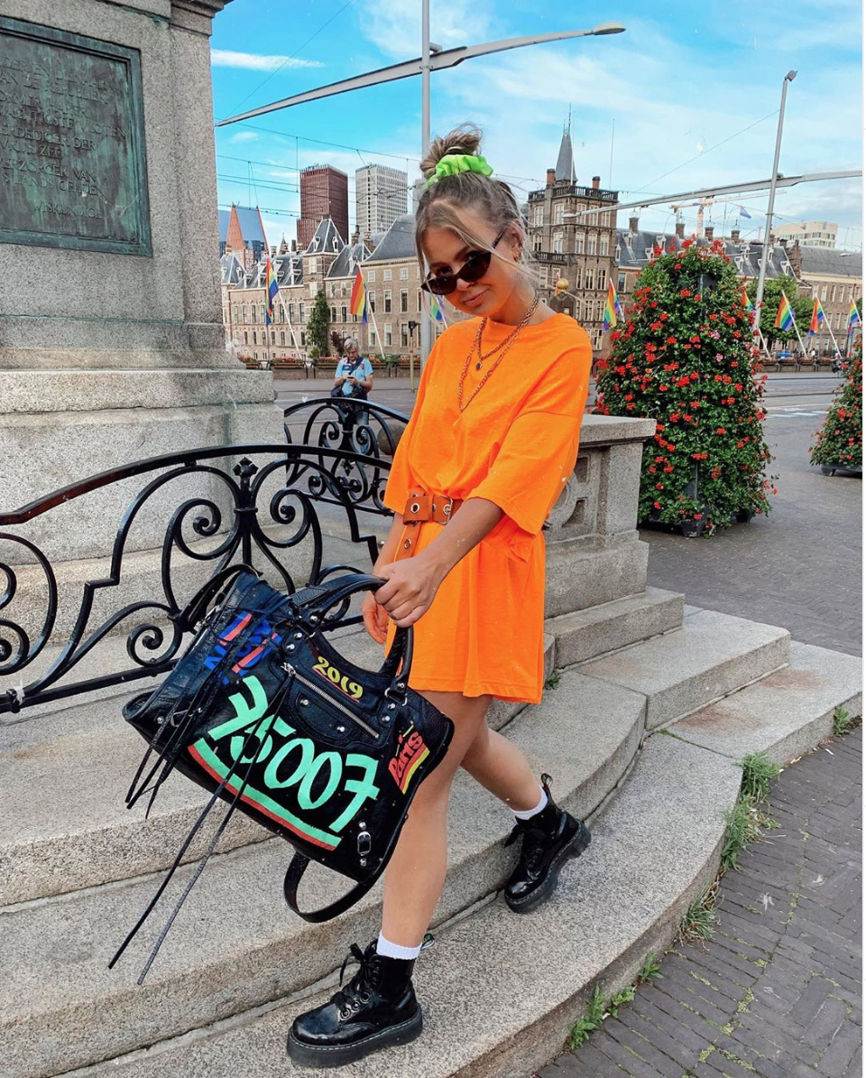 Сумка Balenciaga отлично сочетается с черными грубыми ботинками и оранжевым платьем - футболкой.