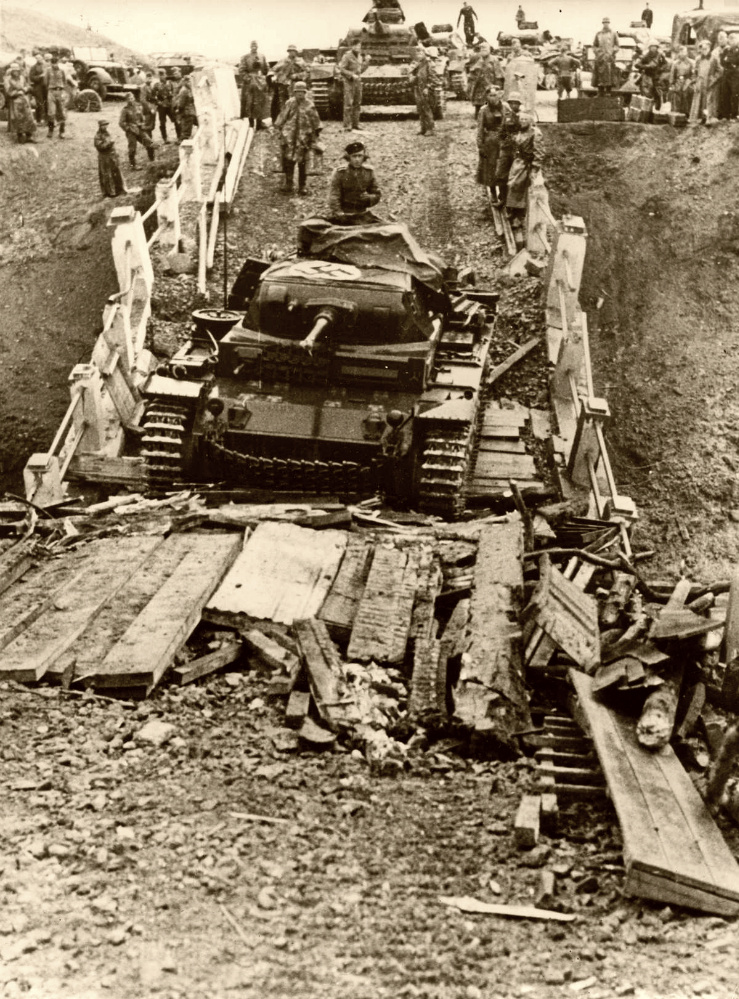 Немецкий средний танк Pz.Kpfw.III Ausf. F преодолевает сильно поврежденный мост. На мосту, чтобы облегчить прохождение войск, разложены деревянные брусья и доски. На втором плане танковая колонна в ожидании марша. Танковая колонна из состава 2-й танковой дивизии. Танк перевооружен 50-мм пушкой KwK 38 вместо штатной 37-мм пушки. 1941 г.