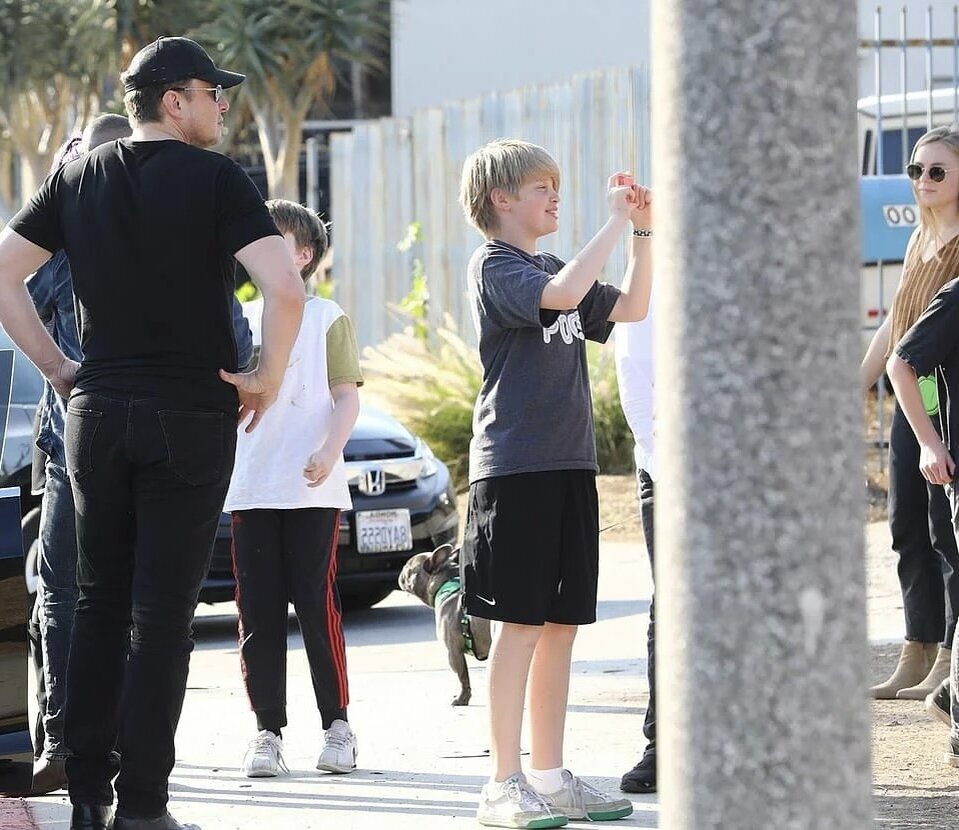 На фото: слева Илон Маск в кепке и очках, в центре его сын Ксавье Маск в серой футболке и чёрных шортах. Фото 2018 года.