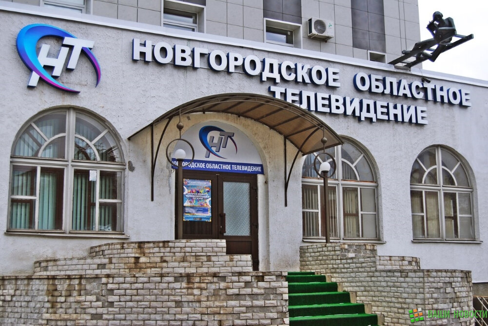 Моё знакомство с НОТ (Новгородское областное телевидение) началось ещё с детства. Я думаю, что многие жители нашего города, так же как и я, включают этот канал только пару раз в день, перед тем как выйти из дома, именно потому, что на нем всегда показывают точную температуру за окном. Я подходил к зданию со стороны парка, с этого ракурса было заметно, что здание покрыто красивой облицовочной плиткой, но только со стороны улицы Большая Московская, а с противоположной стороны до сих пор держится старая, потемневшая краска.

И вот мы вошли внутрь, тут же нас встретили приветливые сотрудники НОТа и позвали пройти на второй этаж. Антураж мигающих лампочек, потертого линолеума, кое-где приклеенного к полу скотчем, создавали ту самую родную атмосферу квартиры бабушки, которая не видела ремонта веками. Там нас встретили удивленные монтажеры, видимо не ожидали, собственно многие из нас также оказались растерянными, ведь лично я ожидал увидеть молодых специалистов, работающих на продвинутом оборудовании, но там нас ждали типичные усатенькие мужчины, ответственно выполняющие свою работу.

На следующем этаже нас ждала аппаратная комната, в которой специально обученный сотрудник следил за вещанием канала. В коридоре одиноко пылилась списанная мебель, видимо ее вынесли с верхних этажей.

Мы будто шли по карьерной лестнице, ведь с каждым этажом становилось все красивее, новее, современнее. Далее мы пришли к кабинетам местных интернет и радио изданий. Их работники были более приветливы, рассказали о себе и о своем деле.
Ускорив шаг, мы двинулись дальше, ведь самое интересное ждало нас выше, там была заветная комната, в которой снимают все передачи НОТа, ведут прямые эфиры и общаются с приглашенными гостями. Это помещение казалось тесным, из-за своей насыщенности разным оборудованием и декорациями, но оно было однозначно из 21 века.

Экскурсия закончилась, и мы поехали вниз на мелодично подпевающем нашим новым впечатлением лифте. А говорят, машины времени не существует…
