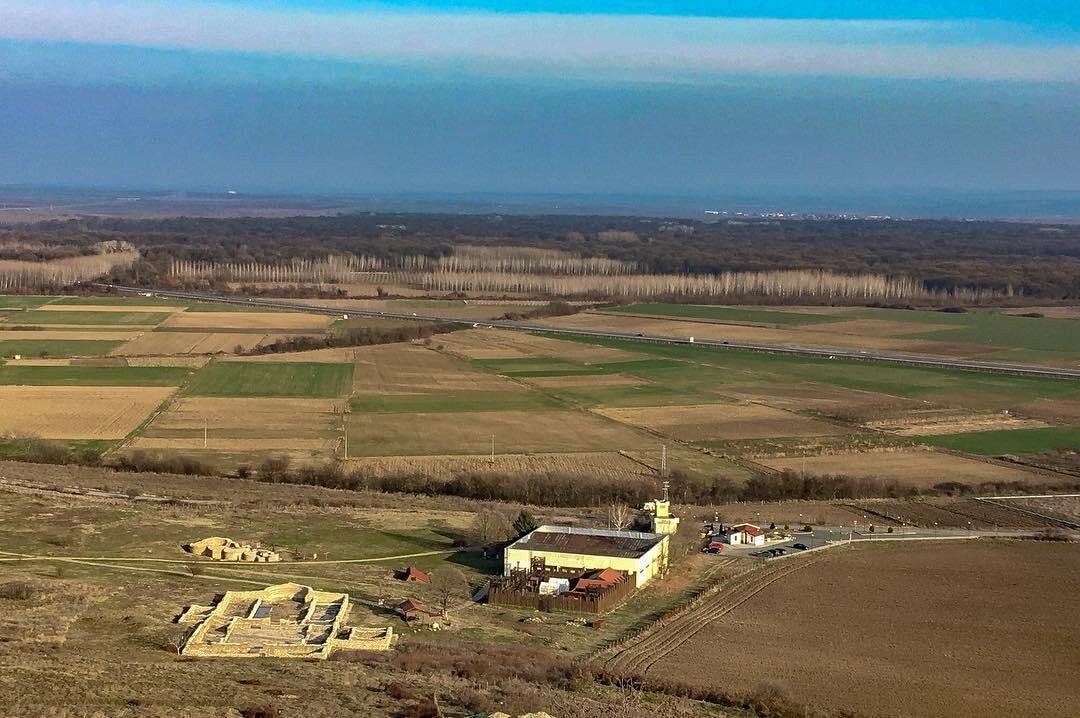 С веришины открывается вид на раскопки. Ближе к нам - руины раннехристианской базилики, где были найдены хорошо сохранившиеся мозаичные полы. Они сейчас выставлены в музее археологического резервата "Тракийский и античный град Кабиле", на фото это желтое строение. А слева за базиликой виднеется небольшая римская терма.