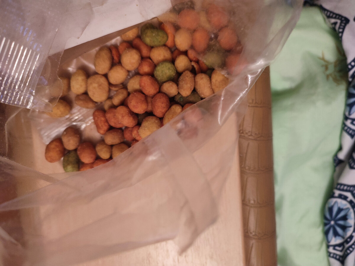 Разноцветные корочки для арахиса<br>
<br>

Вкусные и относительно недорогие арахисы могут быстро приесться. Вот и придумали креативные технологи как их съесть больше).
Арахис в оболочке или как еще называют "в корочке" спрятан от людских глаз в разноцветных шариках, приправленных вкусовыми добавками от нежной сметаны до жгучего васаби или хрена.
Цвет оболочки подобран к специям:

вкус креветок ассоциируется с морем или цветом самой креветки. Поэтому производители окрашивают в нежно коралловый или голубой цвет;
к сырному вкусу соответствуют оттенки желтого;
зеленая оболочка зрительно напоминает цвет горчицы васаби;
светло серый оттенок напоминает цвет хрена;
вкус "салями" или "бекон" - терракотовая оболочка как и сам цвет колбасы
В составе корочки самые обыкновенные ингредиенты:

Мука, кукурузный крахмал (крахмал добавляется, чтобы оболочка была хрустящей);
Соль, сахар;
Вкусовая ароматическая добавка;
Стабилизатор Е1422 - крахмал, необходимый для хранения мучных изделий (в большинстве стран считается безопасная для здоровья пищевая добавка).
Как это делается?

У каждого изготовителя арахиса в оболочке своя технология нанесения глазури на арахис.

Из больших мешков арахис высыпают в бункер и везут по конвейеру в духовку.

в духовых шкафах обжаривают в растительном масле либо нагревают горячим воздухом
в зависимости от технологии и в каком виде подается арахис глазурь наносят путем равномерной присыпки в барабанах, где арахис вертится как в центрифуге белье, периодически брызгая водой для лучшего прилипания корочки
затем арахис охлаждают до температуры окружающей среды и отправляют нам потребителям лакомиться разноцветными вкусняшками