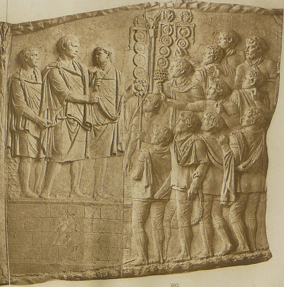 Траян обращается с речью к воинам. Император является наиболее часто изображаемым персонажем украшающих колонну Траяна рельефов. В общей сложности он изображен на ней 59 раз