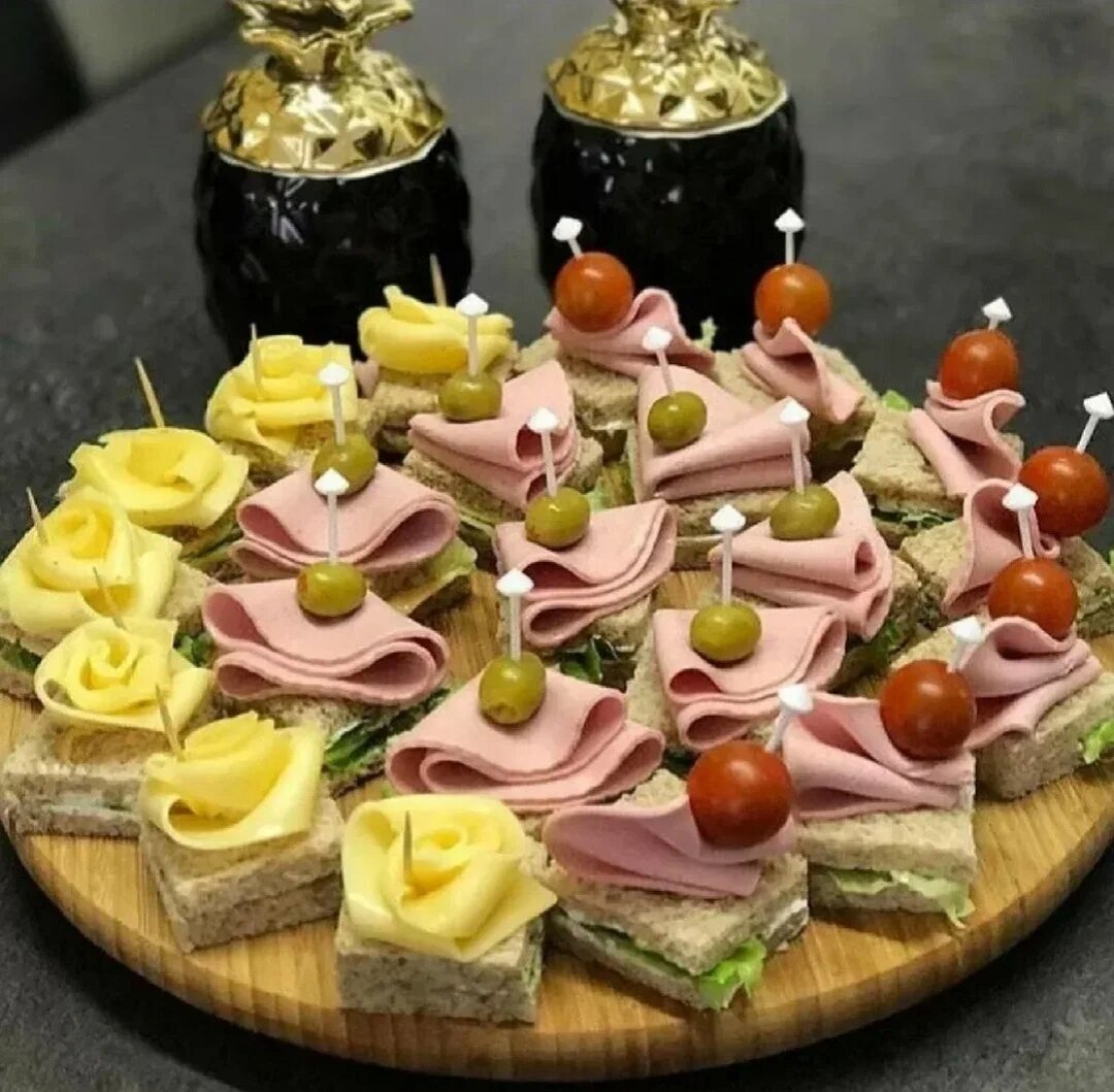 Расположите на круглой деревянной доске канапе: первый слой ломтики хлеба мягким сыром, листья салата, затем снова хлеб, далее украсьте сыром, колбасой или ветчиной, черри и оливки. 