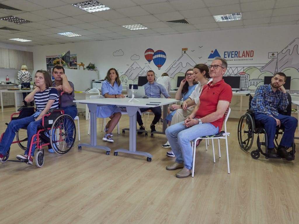 Тифлокомментарий: Одна из встреч в инклюзивном коворкинге Everland — люди с разной инвалидностью (кто-то из них передвигается на коляске) сидят вместе за столом и слушают спикера, который не попал на фото.