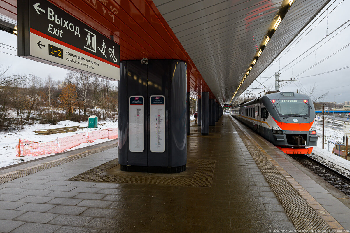 
Станция "Пенягино" - это очень важная станция на западном участке линии МЦД2. Раньше здесь никакой остановки не было, ближайшая остановка электрички была к Красногорске, это станция "Павшино". Однако в начале 2000-х пойму реки Москвы в этом месте начали активно застраивать многоэтажками. Никакой транспортной инфраструктуры здесь не создали, просто застроили весь клочок земли высотками, которые стоят настолько близко, что ни о каком комфортном проживании тут нет и речи. В 2014 году этому району даже присвоили антипремию выставки "Строительная неделя в Московской области" за худшую архитектуру. Вообще в рандере этот район называется "Павшинское гетто". Здесь самая недорогая недвижимость в ближайшем Подмосковье. Все, кто искал себе недорогое жилье в пригороде обязательно натыкался на объявления о продаже квартир тут, некоторые даже ездили смотреть, но ни один человек, которого я знаю, не стал покупать здесь квартиру... Первое время, как я уже говорил, с транспортной доступностью тут было плохо. Сразу же, как начали продаваться квартиры, встало намертво Волоколамское шоссе, выезд из Красногорска превратился в ад. Потом была построена станция метро "Мякинино" но она находится на другом берегу реки, только спустя 5 лет додумались соединить район с метро, перебросив пешеходный вантовый мост. Следующим этапом была организация развязки в месте пересечения Волоколамского шоссе и Павшинской улицы - это в какой то степени решило проблему пробок. И вот, наконец, спустя более чем 10 лет после того, как на этом месте вырос огромный жилой массив, приняли решение о строительстве железнодорожной станции. Это очень правильное, но, как всегда бывает у РЖД, сильно запоздавшее решение. Но, как говорится, лучше поздно...