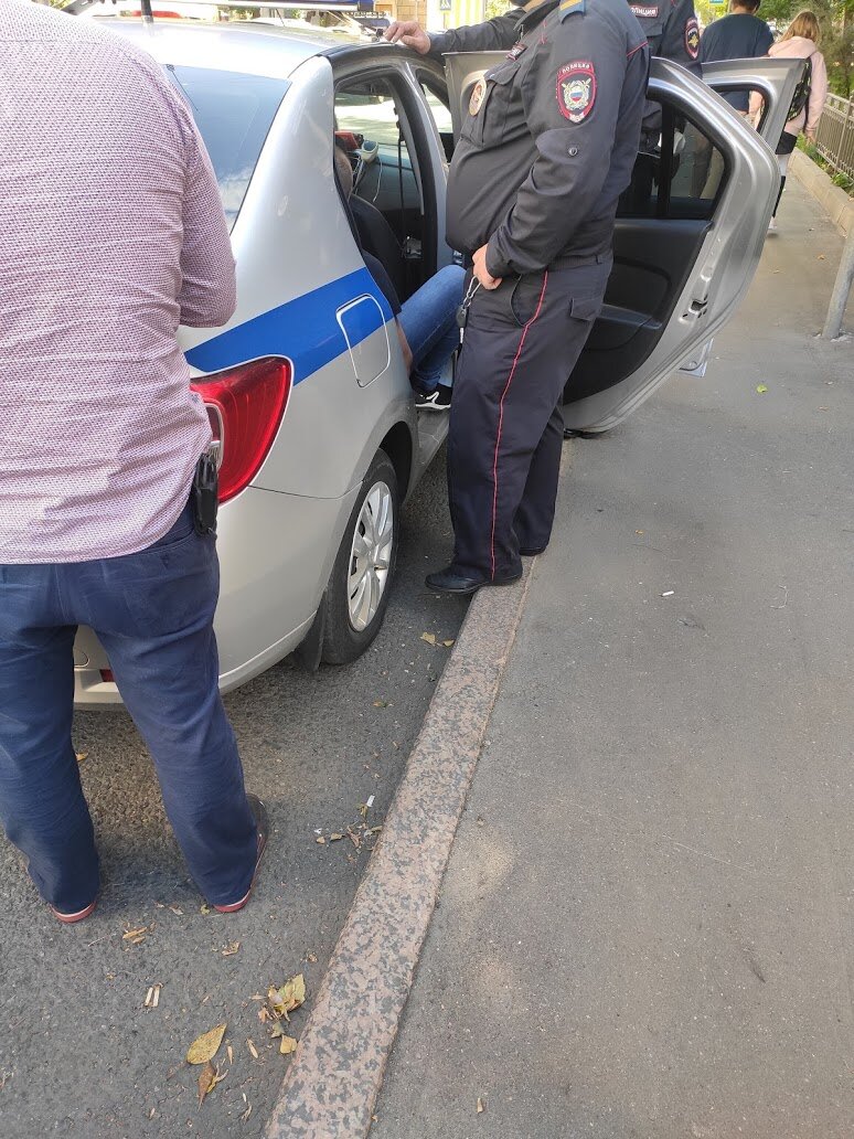 Подозреваемый сидит в машине. Сзади машины оперативник.