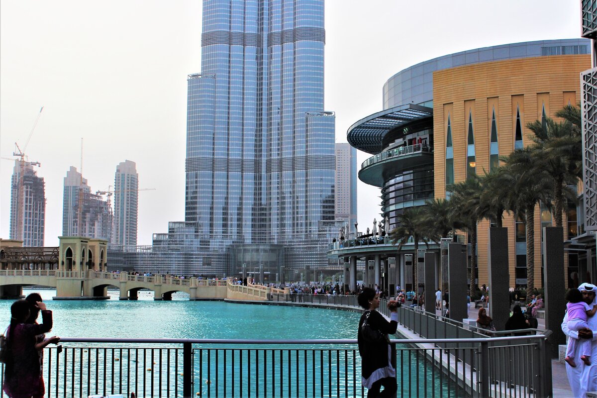 В средине фотографии башня-игла, справа вход в Дубаи Молл, мост перекинут через бассейн, где в носи поют и сверкают огнями поющие фонтаны