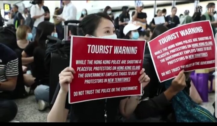 Девушка протестует в аэропорту Гонконга. Текст плаката: "ПРЕДУПРЕЖДЕНИЕ ТУРИСТАМ. Пока полиция Гонконга стреляет в мирных протестующих на острове Гонконг, правительство нанимает головорезов, чтобы они избивали протестующих на новых территориях Не доверяйте полиции или правительству.".