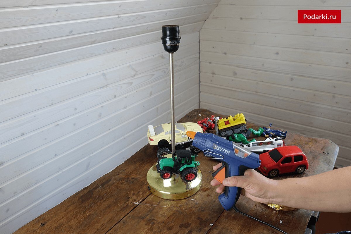 Среди игрушек, которые вы выберете для работы, должно желательно использовать несколько крупных машинок — они станут хорошей основой композиции. Одну из них приклейте так, чтобы она стояла четырьмя колесами на основание настольной лампы (для крепости конструкции). 