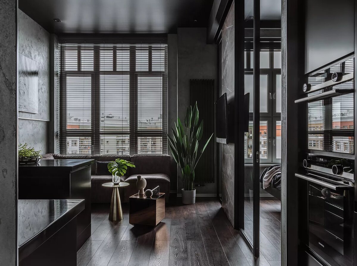 Массивный угловой диван отнесли к окну. Его брутальность разбавили элегантными кофейными столиками и живыми растениями. Перегородка со спальней почти полностью стеклянная, исключение сделали только для ТВ-зоны.