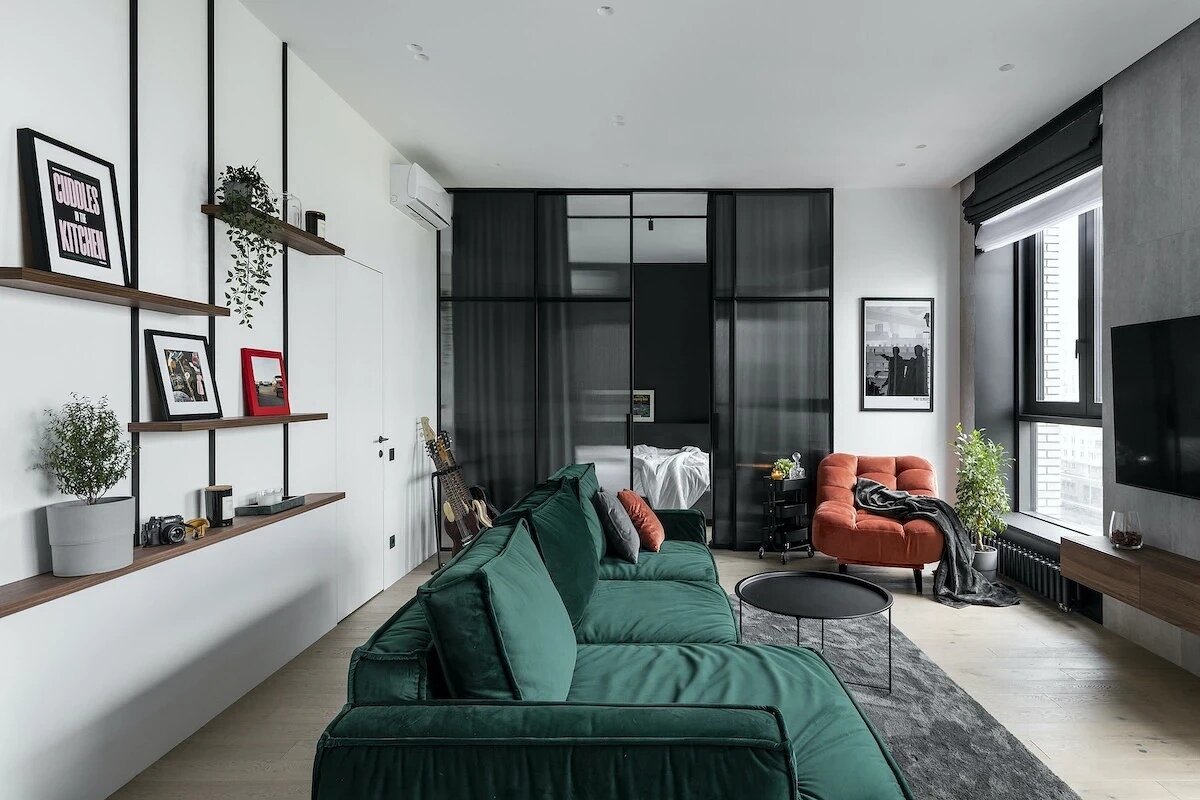 Диван поставили спинкой к проходу, тем самым очертив границы гостиной. За ним повесили полки для декора и живых растений. Мягкую группу дополнили креслом в терракотовой обивке. Панорамные окна завесили римскими шторами в тон откосам и подоконникам.