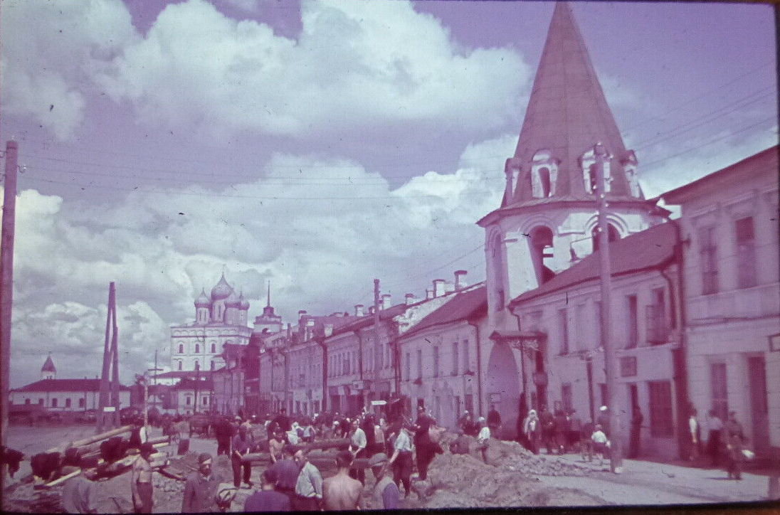 Слева церковь Михаила Архангела. Недавно попала в Юнеско. Вдали Троицкий собор. На первом плане часть улицы капитально перекопана. Пленных не вижу, скорее вольнонаемные и ТОДТ, возможно ж.д. батальон укладывает ж.д. рельсы к мосту Красной Армии (Ольгинскому). По тротуару ходят гражданские и дети. Справа около церкви должна быть лавка с книгами. Датировка -скорее всего лето 1942 года.