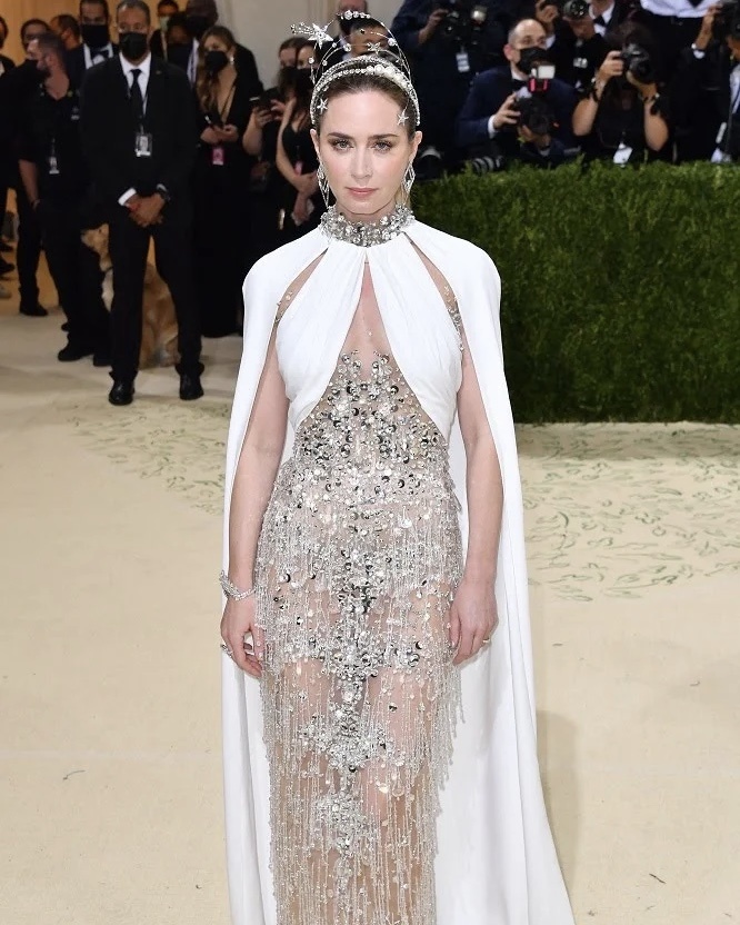 Эмили Блант была в MIU MIU на мероприятии MET Gala 2021, посвященном открытию выставки “In America: A Lexicon of Fashion” (В Америке: Лексикон моды) 14 сентября 2021 года в Нью-Йорке.
Актриса была одета в прозрачное платье-колонну Miu Miu с корсетом из тюля телесного цвета с драпировкой из белого шелка, украшенное вышивкой из кристаллов, бахромой из кристаллов и деталей в виде звезд по всей поверхности, дополнив образ босоножками с кристаллами и повязкой с декором из жемчуга и элементов в виде звезд.