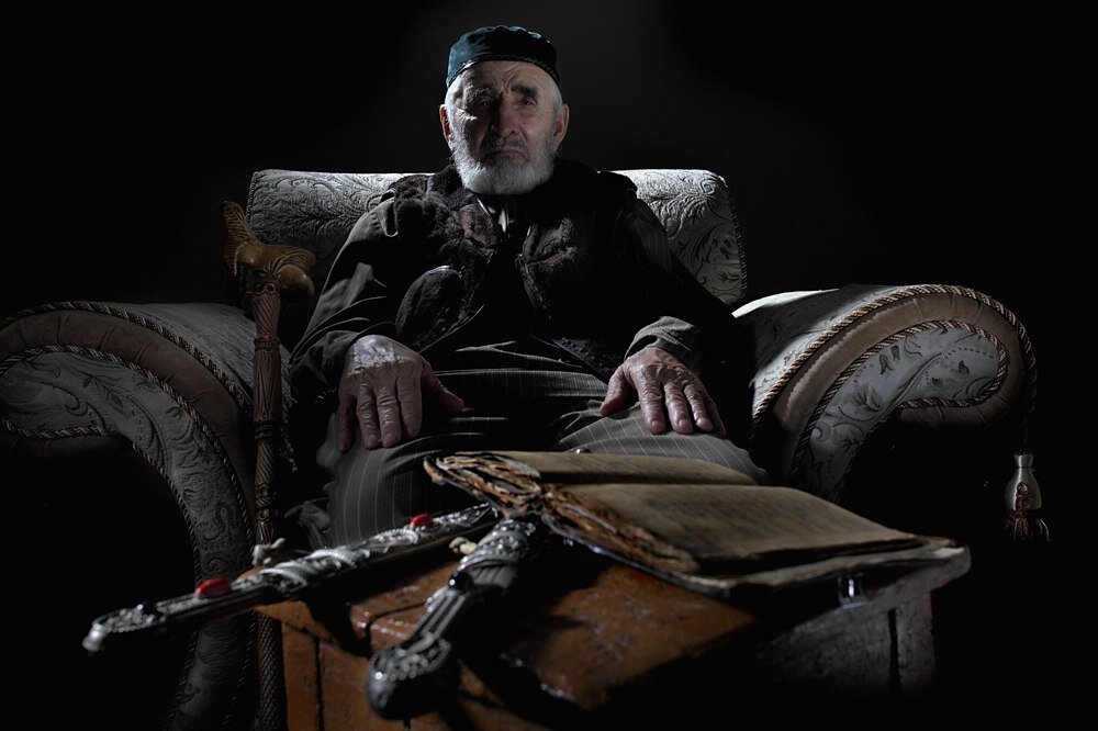 © Дмитрий Беляков. Иса Хашиев, Ингушетия, 80 лет. Позирует с сохраненными в ссылке древним Кораном и кинжалами его отца и деда. «Этому Корану больше ста лет. И вот этому кинжалу тоже. Он моему отцу принадлежал. Когда ему 16 лет было, ему купили. А сколько второму кинжалу, я даже предположить не могу. Он дедов. Мы все это в матрасах спрятали, когда нас выселяли. Что сказать? Плохо было, когда ехали. Условия в пути были ужасные. Когда прибыли на место, всю ночь нас везли на санях по степи в колхоз какой-то. Вьюга, снег, ночь, холод. Моя младшая сестренка тогда сильно заболела и умерла. Ей всего несколько месяцев было. Отчего умерла? Замучилась… Взрослые-то замучились. А меня в пути угораздило с саней этих свалиться. Упал, очнулся в сугробе. Испугался ужасно. Мать почувствовала, что меня нет, крик подняла. Нашли меня, а то замерз бы… Что еще помню? Что нам вообще-то повезло: мы-то в русский колхоз попали, там были русские поселки. Там к нам по-человечески отнеслись. Если было зерно на складе, то давали. И молоко давали. А казахи… они, конечно, считаются мусульманами, но, вообще-то, они к нам, мусульманам, относились хуже, чем русские. Если бы не русские, наши бы не выжили в тех условиях. Спасли они нас тогда».
