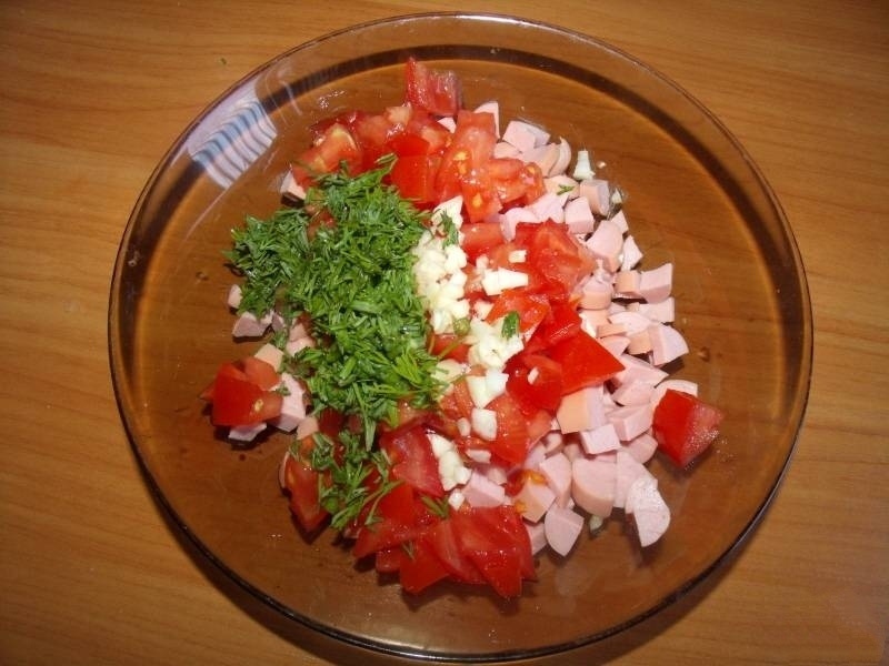 Помидор так же нарезаем кубиками, зелень мелко шинкуем и добавляем в тарелку к сосискам. Выдавливаем чеснок через чесночницу.