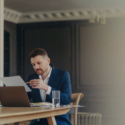    Handsome business executive in dark blue suit sitting at his work desk in light colored office working on data presented on A4 sheet, holding pencil while being serious and concentrated fabrica