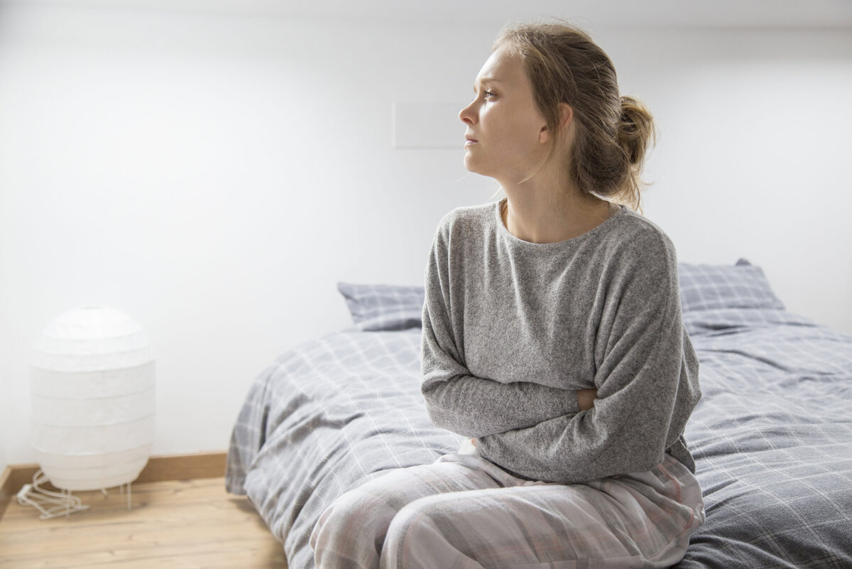    Tired sick woman in grey homewear sitting on bed, keeping hands on stomach, suffering from pain, looking aside. Illness, stomach ache concept