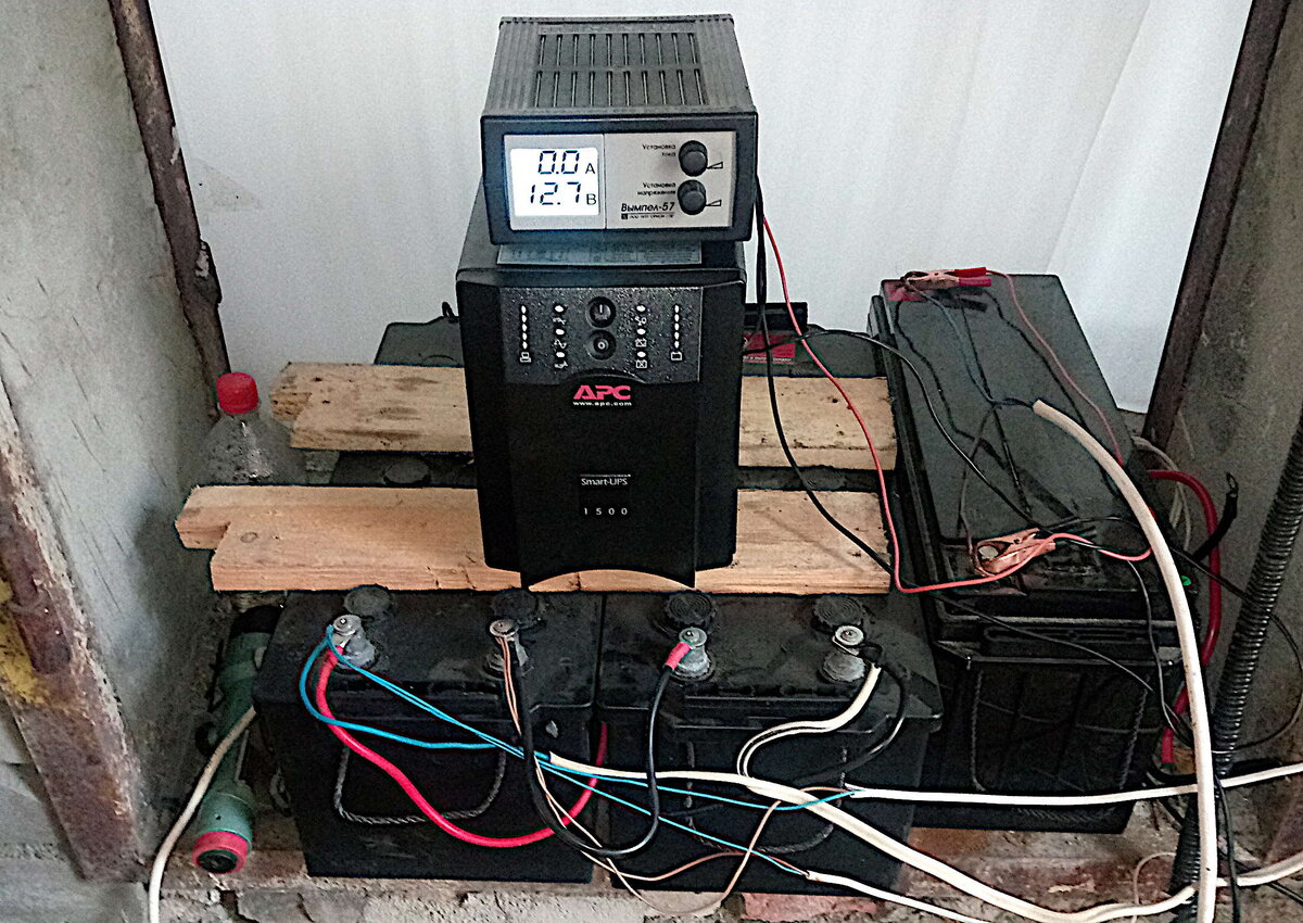 Фото автора. Сверху зарядное на 12 В - подключено к AGM 150, туда-же подключен ветряк. Ток показывает только в режиме заряда от 220 В, сейчас всё выключено. Ниже источник 220 В на 1500 ВА - 950Вт. Под ним два аккумулятора по 190 АЧ. К одному из них на 12В  подключена местная подсветка светодиодная и к обоим на 24В толстым белым проводом - регулятор заряда солнечной батареи и туда-же APC. 
