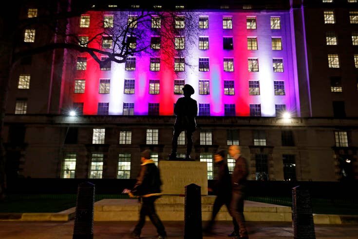 Пешеходы проходят перед зданием Министерства обороны на Уайтхолле, освещенном красными, белыми и синими огнями в центре Лондона