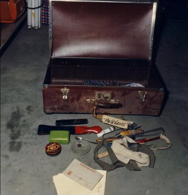 На этом фото видны следующие предметы: чемодан с биркой вещ.дока на его ручке, три галстука — один полосатый в чемодане, два других, белый и серый, лежат на полу, нож, кисть, стеклянная тарелка с пуговицей и ложка, крем для обуви марки KIWI, зеленая мыльница, зубная паста и щетка, помазок, ножницы в «ножнах» в рабочем состоянии, от вторых ножниц, сломанных, выглядывает только ручка с кольцом, карандаши, вешалки, опасная бритва в футляре и рядом правИло для нее, на переднем плане видны конверты с лежащим поверх них письмом-секреткой.