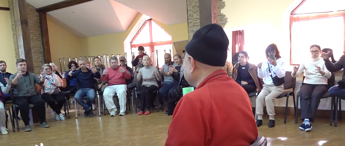 Му Юйчунь сидит в центре. Люди сидят на стульях по периферии зала. Скриншот учебного видео.