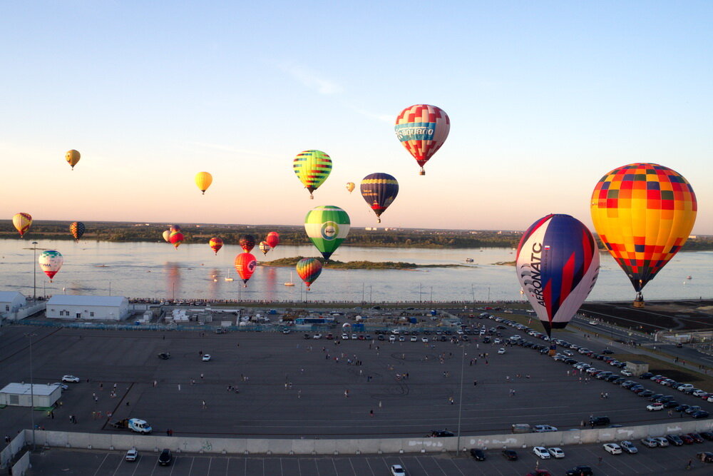 Кажется, что на огромной стоянке перед пустующим стадионом собрался весь город. Все рассматривали аэростаты и определяли самый красивый. Воздушные шары летали над стрелкой и раньше, но впервые из собралось такое количество – 25. Казалось, что места для всех не хватит, шары стояли толкая друг друга, но в итоге все смогли разместиться. Долго ждали метео – юго-западный ветер дул порывами и стихать не хотел, сдался лишь под самый вечер. И тут шары начали подниматься. Один за одним. В течение нескольких минут парковка опустела, и шары стали видны тем, кто гулял по набережным. Они пересекли Волгу и плавно сели на Бору с тем, чтобы буквально послезавтра порадовать горожан ночным свечением. Приходите! Будет интересно!