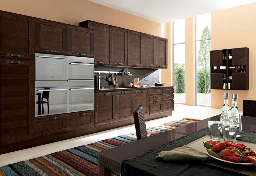 БРУТАЛЬНАЯ ПРЯМАЯ КОРИЧНЕВАЯ КУХНЯ В НЕОКЛАССИЧЕСКОМ СТИЛЕ ДЛИНОЙ 5,4 М. ЛЕВАЯ ЧАСТЬ КУХОННОГО ГАРНИТУРА ОТВЕДЕНА ПОД ПЕНАЛЫ ДЛЯ ВСТРОЕННОЙ ТЕХНИКИ И ХОЗЯЙСТВЕННЫЙ ШКАФ, А В ПРАВОЙ РАСПОЛОЖЕНА РАБОЧАЯ ЗОНА. ПЕРЕПАД ВЫСОТ МЕЖДУ ПЕНАЛАМИ И ВЕРХНИМИ ШКАФАМИ ПРИДАЁТ ОБСТАНОВКЕ ДИНАМИЧНОСТИ. ЧЁРНЫЕ ШИРОКИЕ РУЧКИ ОТЛИЧНО СОЧЕТАЮТСЯ С ТОНИРОВАНЫМИ ФАСАДАМИ ИЗ МАССИВА ДУБА, А ОБЛИЦОВАННАЯ ПЛАСТИКОМ СТОЛЕШНИЦА И СТЕНОВАЯ ПАНЕЛЬ С ДЕКОРОМ ПОД ЛИСТ ЖЕЛЕЗА С РЖАВЧИНОЙ ПРИВНОСИТ В ЭТОТ МУЖСКОЙ ИНТЕРЬЕР НОТКУ ЛОФТА.