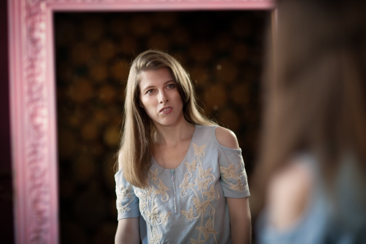 Тифлокомментарий: Светлана стоит перед зеркалом и смотрим на себя, видно только ее отражение до пояса. У девушки распущены волосы, она одета в светло-голубую рубашку с коротким рукавом и открытыми плечами. 