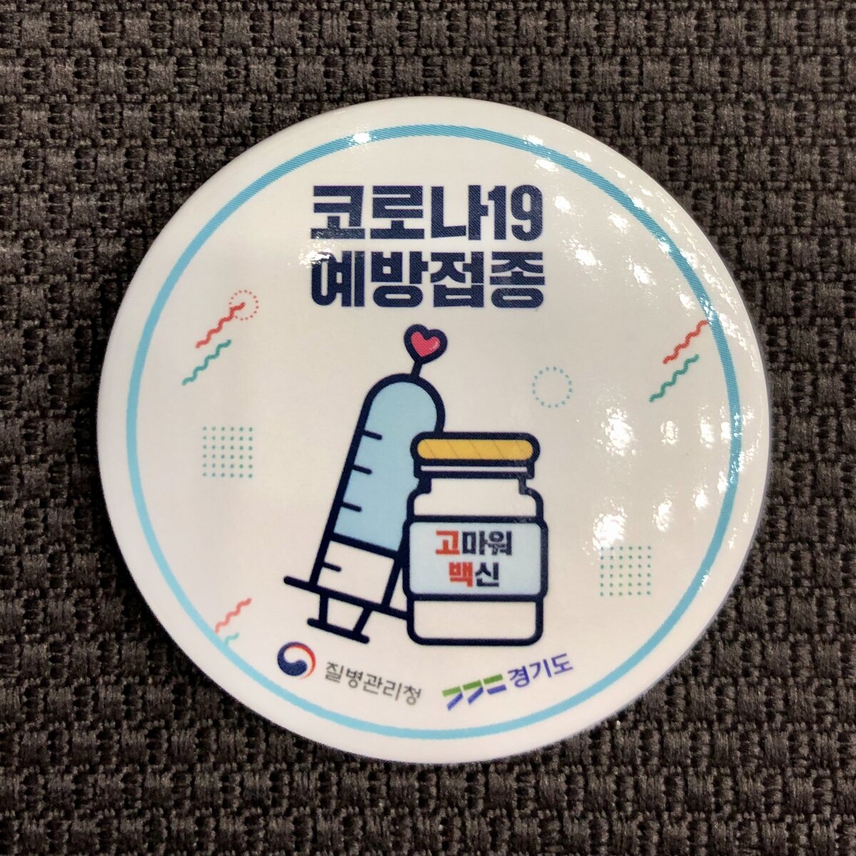 После прививки меня наградили памятным значком. На нём сверху написано ПРОФИЛАКТИЧЕСКАЯ ПРИВИВКА ОТ КОРОНЫ-19. На баночке надпись «Спасибо, вакцина!». Внизу — «Управление контроля заболеваний» и «Кёнгидо».