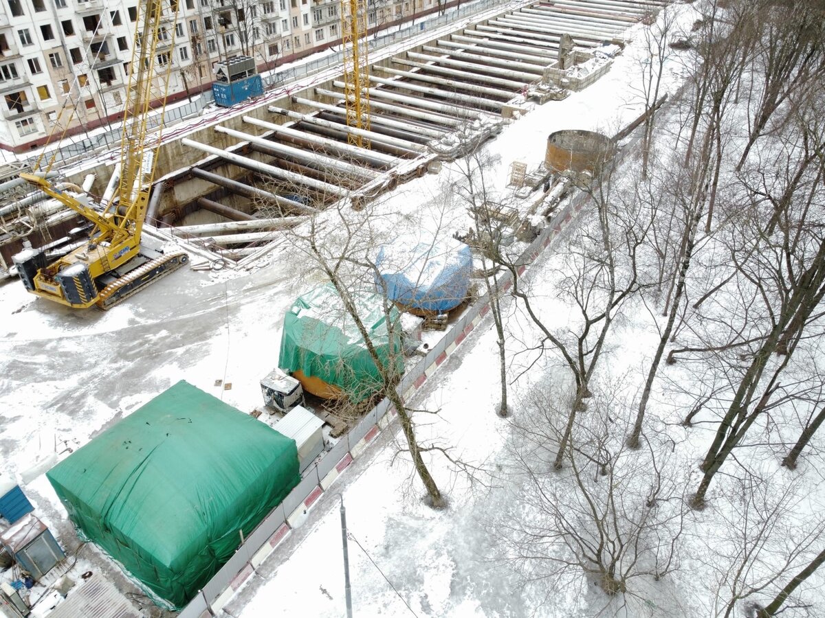 📷 1: Около монтажной камеры в сторону «Бульвара Карбышева» складированы детали головной части второго ТПМК. Часть его зищитовых телег уже спущено в котлован, но три ещё находятся на поверхности. Первый ТПМК накрыт зелёным тентом, бетонный куб, внутри которого находится головная часть, готов. Рядом с котлованом лежат блоки тоннельной обделки, которых хватит для врезки первого щита.