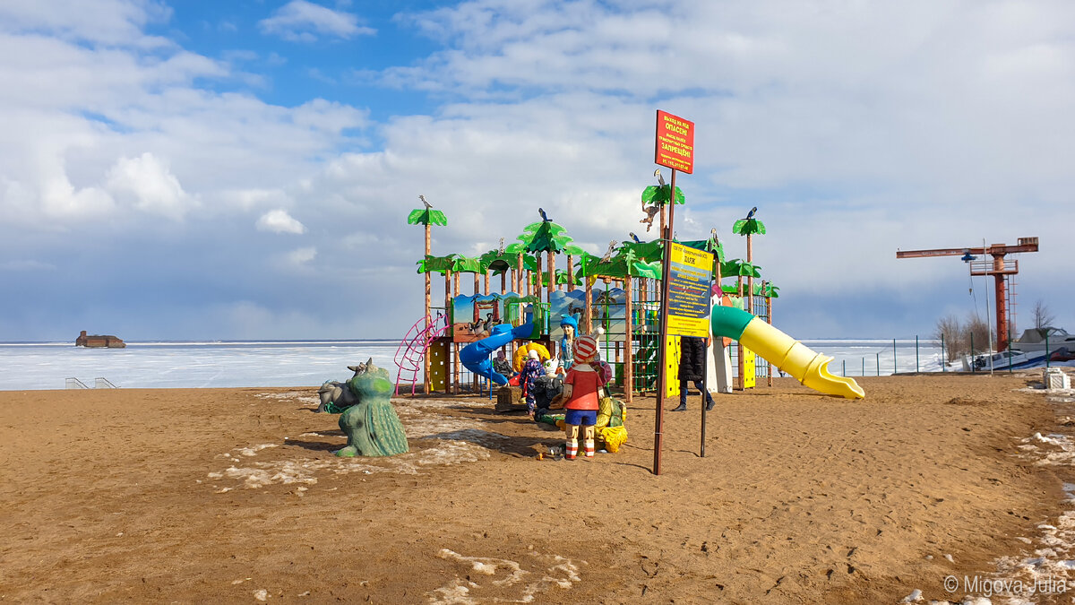 На фоне затянутого льдом залива детская площадка тропического вида выглядит сюрреалистично