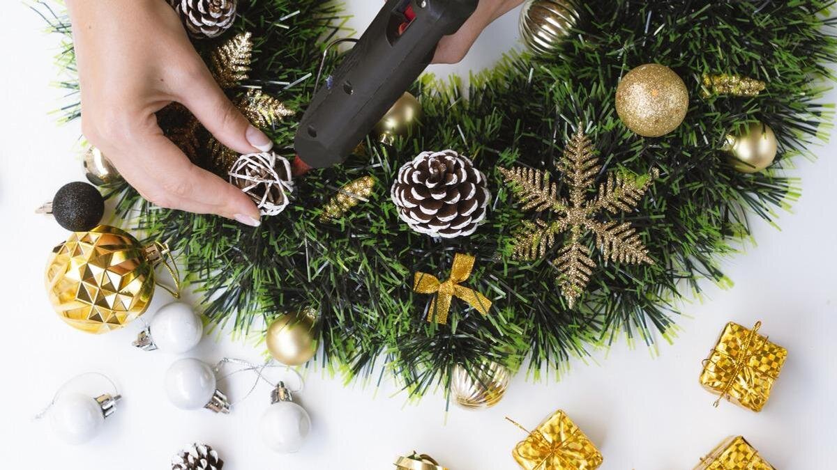    К новогоднему венку из искусственной хвои клеевым пистолетом приклеивают шарик из ротанга. Рядом лежат подарочные коробочки, гирлянда, шишки:Freepik