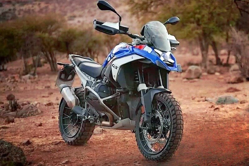 Фотографии из пресс-кита, подготовленного к официальной презентации нового BMW R1300GS 2024 модельного года, просочились в сеть за неделю да даты премьеры. Специально, или случайно, уже не поймёшь, но интерес к премьере новинки они подогрели. Дело в том, что при более-менее том же железе, был радикально изменён дизайн, и кое у кого от взгляда на совершенно симметричный анфас знатно подгорело.
Ушла в прошлое концепция дизайна «Подбитый глаз», когда одна фара была крупнее другой. Сейчас ровно в центре размещается крупная Bi-LED линза дальнего/ближнего света, от которой в разные стороны расходятся диагонали квадрата ходовых огней. Морда стала лаконичнее и компактнее, да и сам мотоцикл похудел и стал более поджарым, как мужчина, наконец-то вставший с дивана и переставший пропускать тренировки по кросс-фиту.

У нового мотора с сухим картером и отдельным маслобаком мощность должна вырасти до 145,5 л.с. при 7750 об/мин, что не выводит мотоцикл, в лидеры класса, но несколько сокращает отставание от лидеров. Момент ожидается 149 Н*м при 6500 об/мин. Ход поршня остался прежним, диаметр цилиндра немного вырос – за его счёт добились увеличения рабочего объёма. Подвески остались прежними по конструкции – «Телелевер» с «Паралевером», но колёсная база немного увеличилась – на 4 миллиметра до 1518 мм.
Из приятных моментов также стоит отметить по-новому проложенные патрубки выхлопа от цилиндров, при падении мотоцикла они не должны страдать. А ещё новые дуги, защищающие сами цилиндры. Похоже, не изменяя своим компоновочным принципам и приоритетам на комфорт и туризм, в BMW получилось добавить немного огня в большого «Гуся», который стал ещё больше, но при этом спортивнее.
Официальная премьера мотоцикла намечена на четверг, 28 сентября 2023.