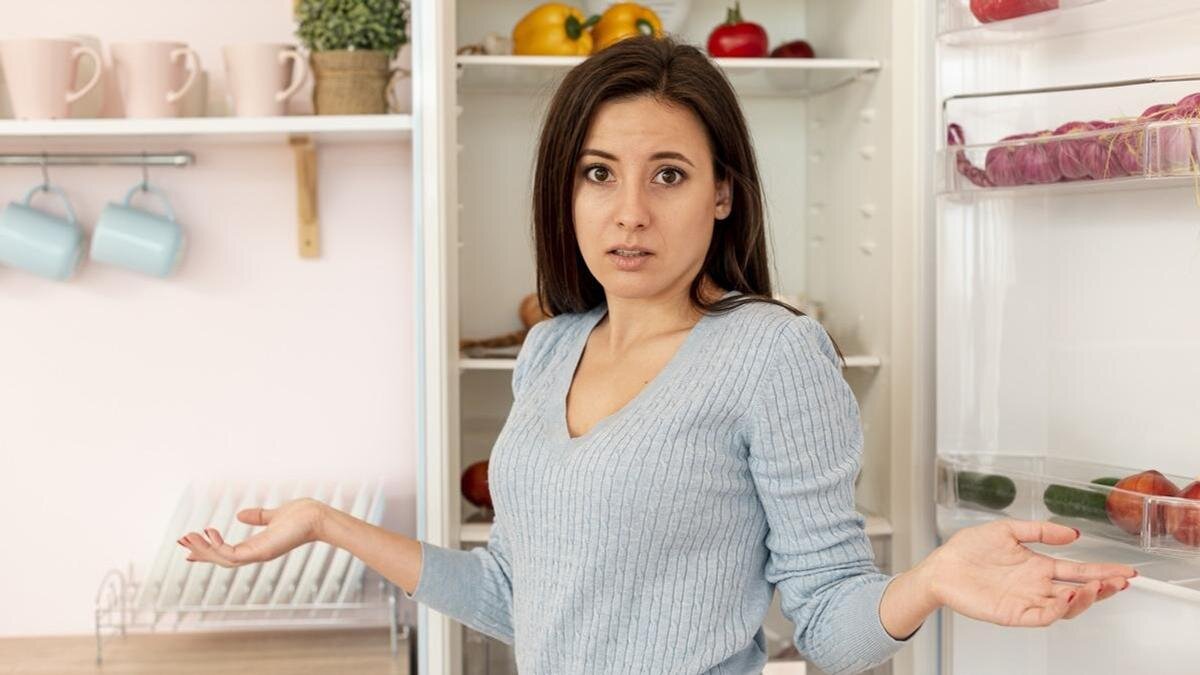    Девушка стоит перед открытым холодильником с продуктами и разводит руками:Freepik