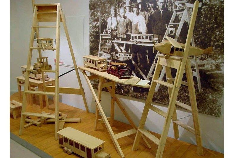 Продукция LEGO образца 1932 года в музее компании. На заднем плане — рекламный снимок LEGO: весь состав мастерской и товары.