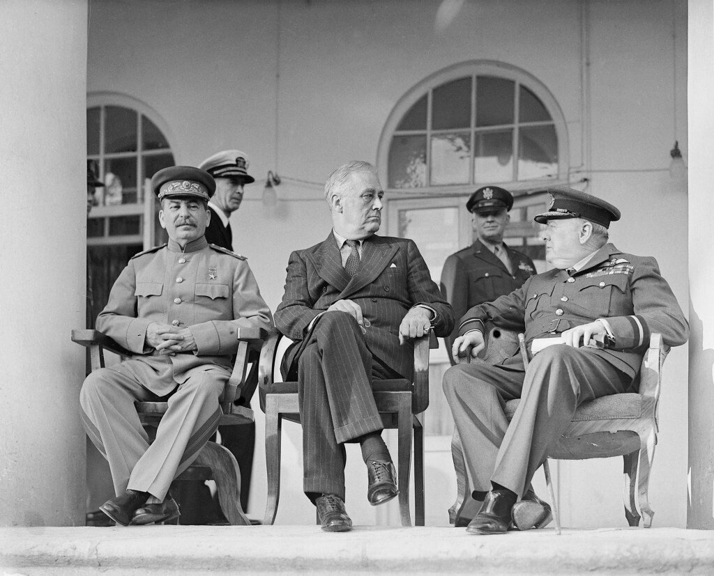 Иосиф Сталин, Франклин Рузвельт и Уинстон Черчилль сидят на стульях на веранде советского посольства. На заднем плане - помощники президента США. Иран. Тегеран. 29 ноября 1943 года. Имперский военный музей (Великобритания). А20710