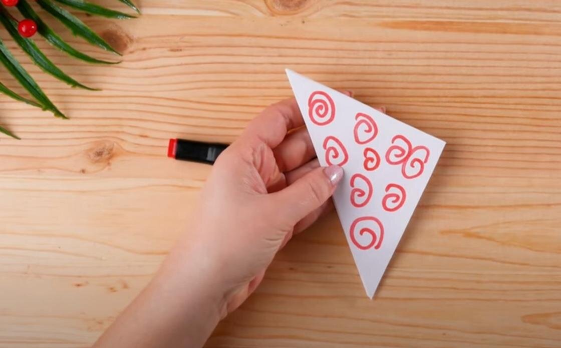    В руке держат треугольную бумажну. хлопушку с нарисованными красными завитушками. Рядом лежит красный маркер и ветка искусственной зелени:YouTube/ОРИГАМИ И НЕ ТОЛЬКО