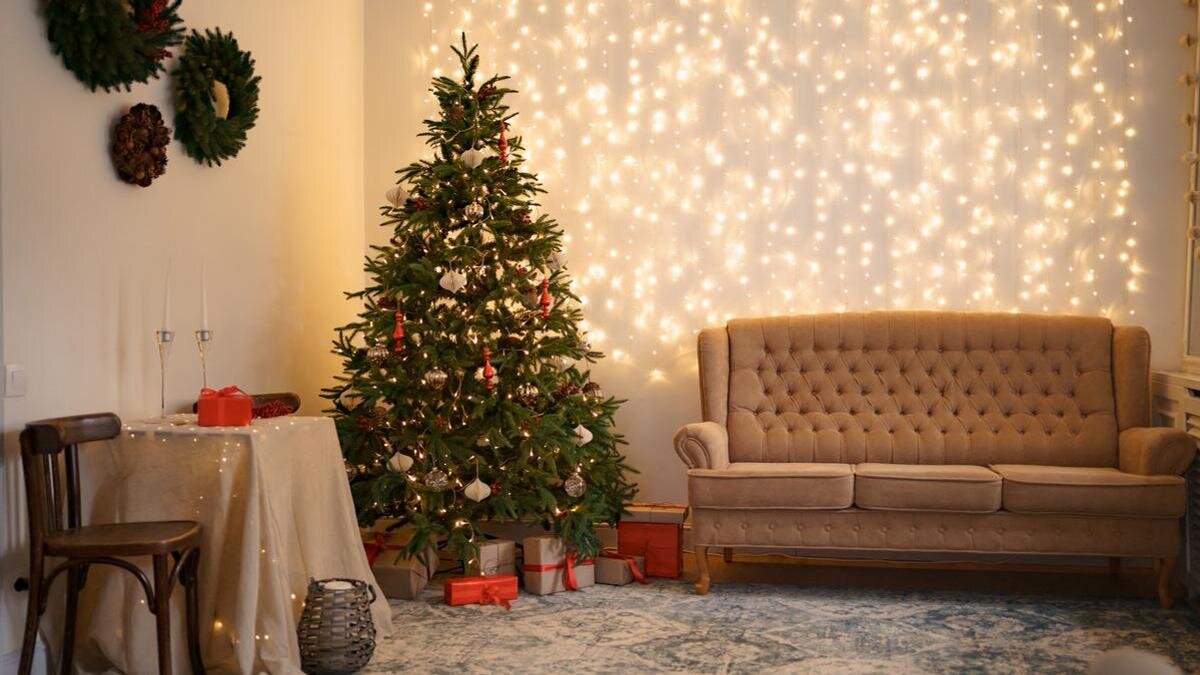    Новогодняя елка стоит на подставке в углу дома. Под стеной стоит диван, стул, столик, а на стене висят новогодние венки:Freepik