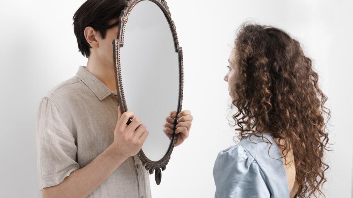    Парень держит в руках небольшое зеркало в раме и закрыл им лицо. Девушка стоит напротив и смотрит в зеркало:Freepik