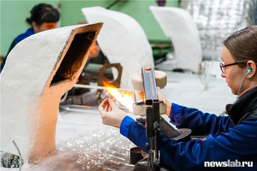 В шумном помещении в наушниках сидят стеклодувы, на их рабочем месте — керосиновая горелка. Стеклодув берет заготовку и начинает крутить ее над пламенем горелки. Стекло становится мягким, и тогда уже можно выдуть шар или игрушку в форме рыбки, например