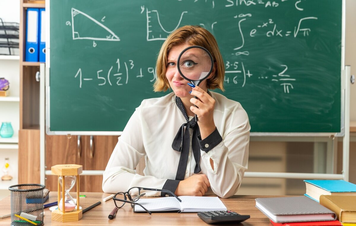 impressed-young-female-teacher-sits-at-table-with-school-tools-with-magnifier-in- Используется для иллюстрации