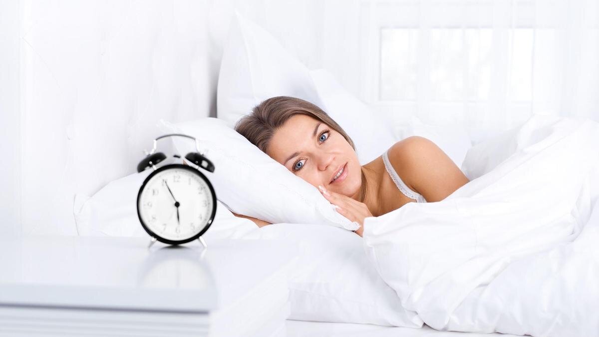    Девушка лежит в кровати, будильник стоит на тумбочке:Freepik