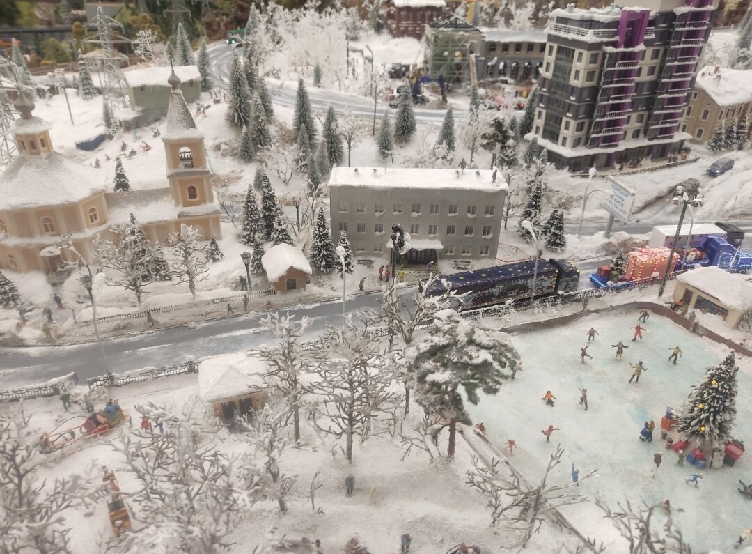 Зимний город. По дороге ездят машины, в окнах мигает свет, а на катке горит ёлка.